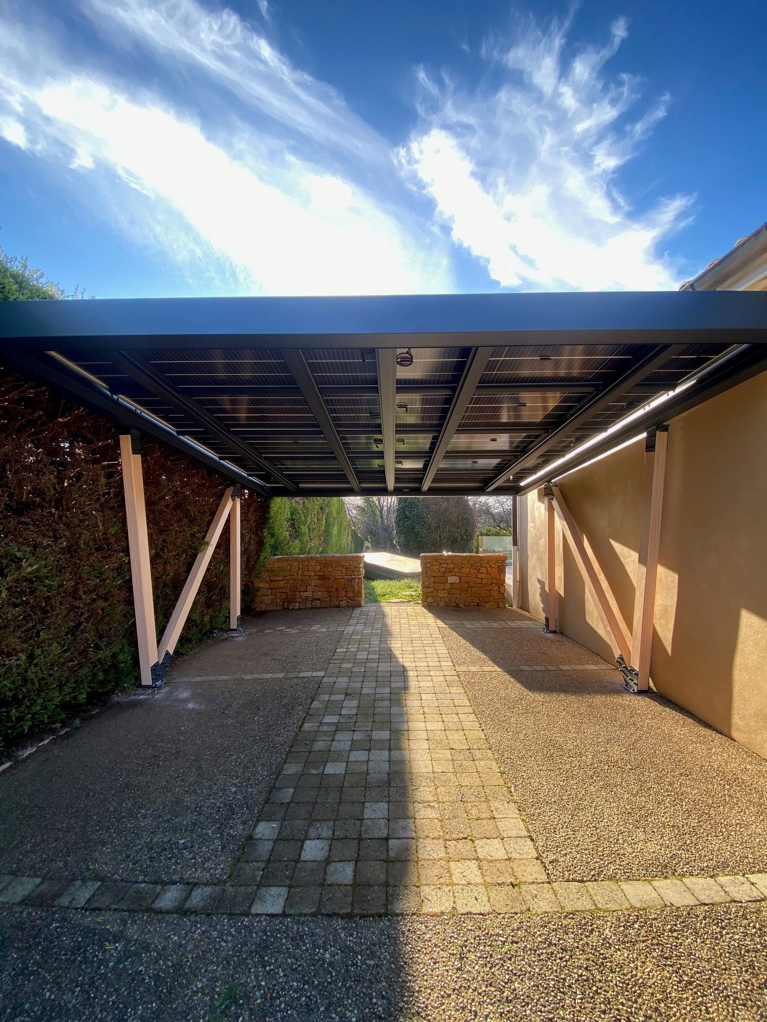 Carport solaire allongé couvrant une allée pavée entre une haie et un mur, vu dans l'axe