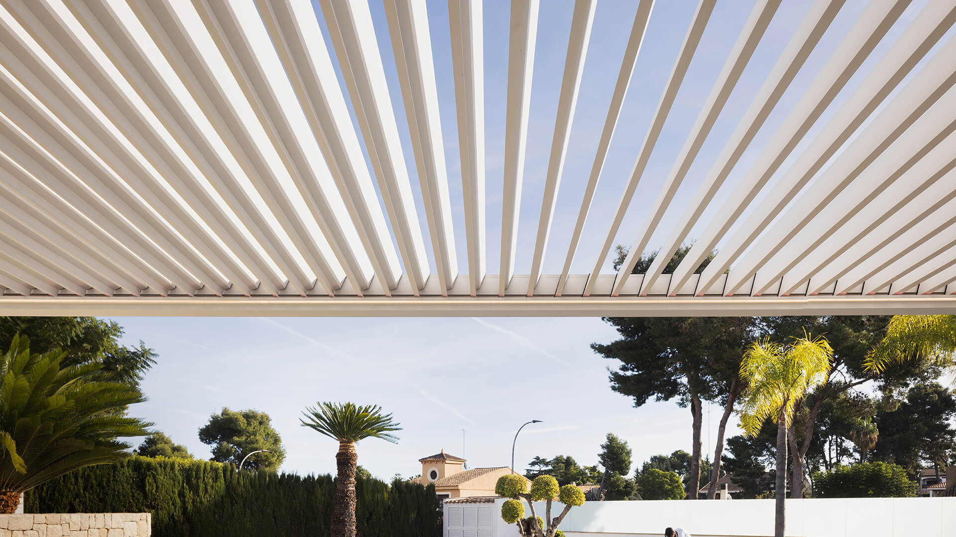Lames orientables ouvertes d'une pergola bioclimatique blanche vues de dessous, sur fond de ciel et de palmiers
