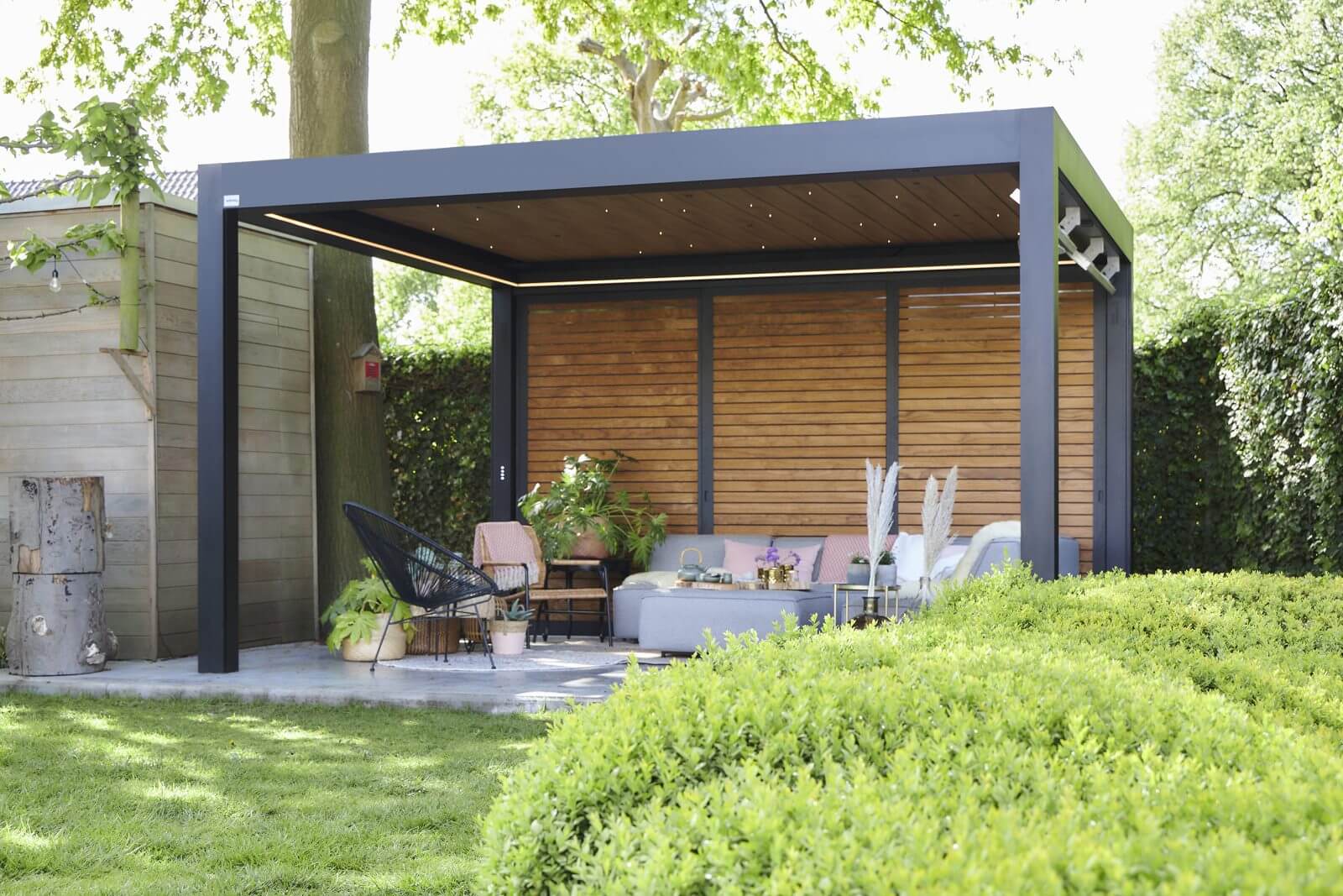 Pergola anthracite au plafond d'aspect bois avec panneau de fond en bois, abritant un salon de jardin