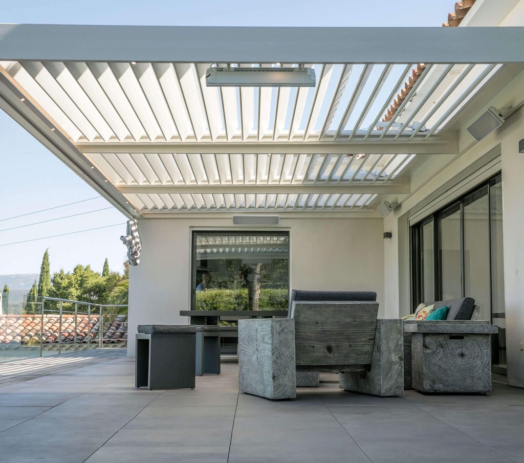 Une terrasse équipée face aux collines Pergola bioclimatique blanche adossée, équipée sous ses lames, au-dessus d'une terrasse meublée avec vue sur les collines