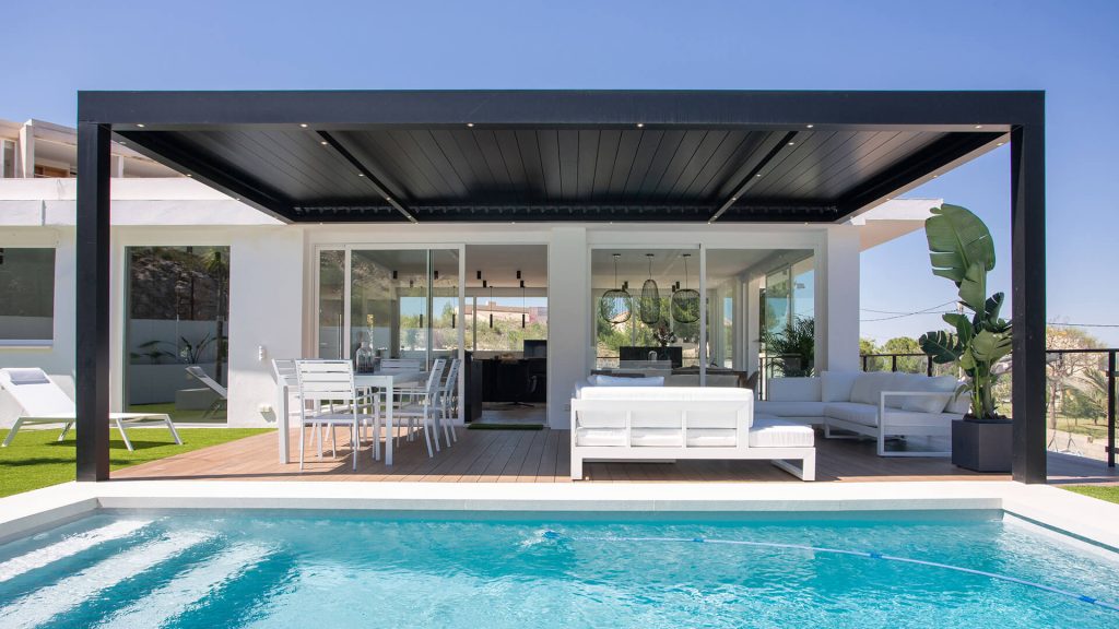 Un salon d'été au bord de la piscine Pergola bioclimatique anthracite couvrant la terrasse d'une villa blanche, au bord d'une piscine