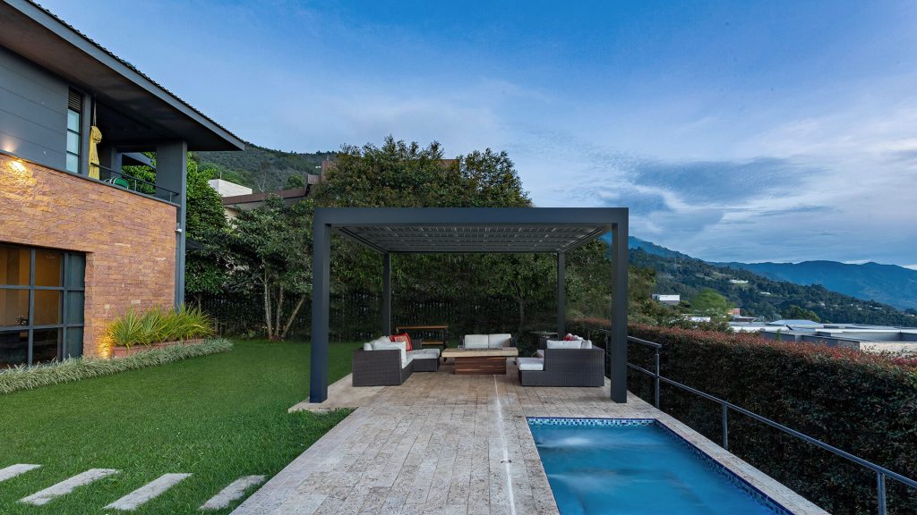 Un salon extérieur face aux montagnes Pergola bioclimatique anthracite indépendante abritant un salon de jardin, près d'un bassin et d'une maison en briques