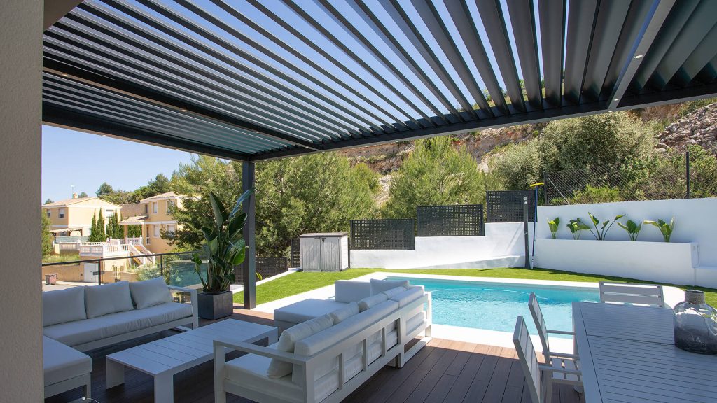 Un coin détente à l'ombre près de la piscine Pergola bioclimatique anthracite aux lames ouvertes abritant un salon blanc, avec vue sur la piscine et le jardin