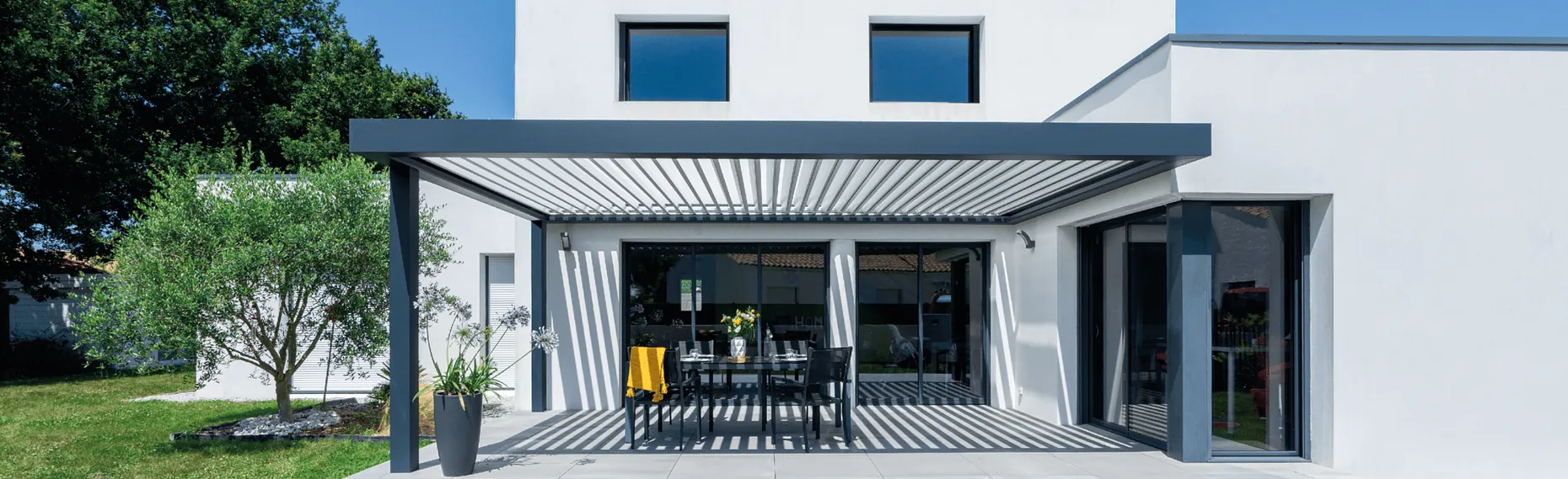 Pergola bioclimatique adossée aux lames ouvertes, abritant une table de repas sur la terrasse d'une maison contemporaine