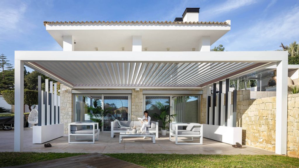 Un salon extérieur devant la villa Pergola bioclimatique blanche aux lames ouvertes abritant un salon de jardin devant une villa aux murs de pierre