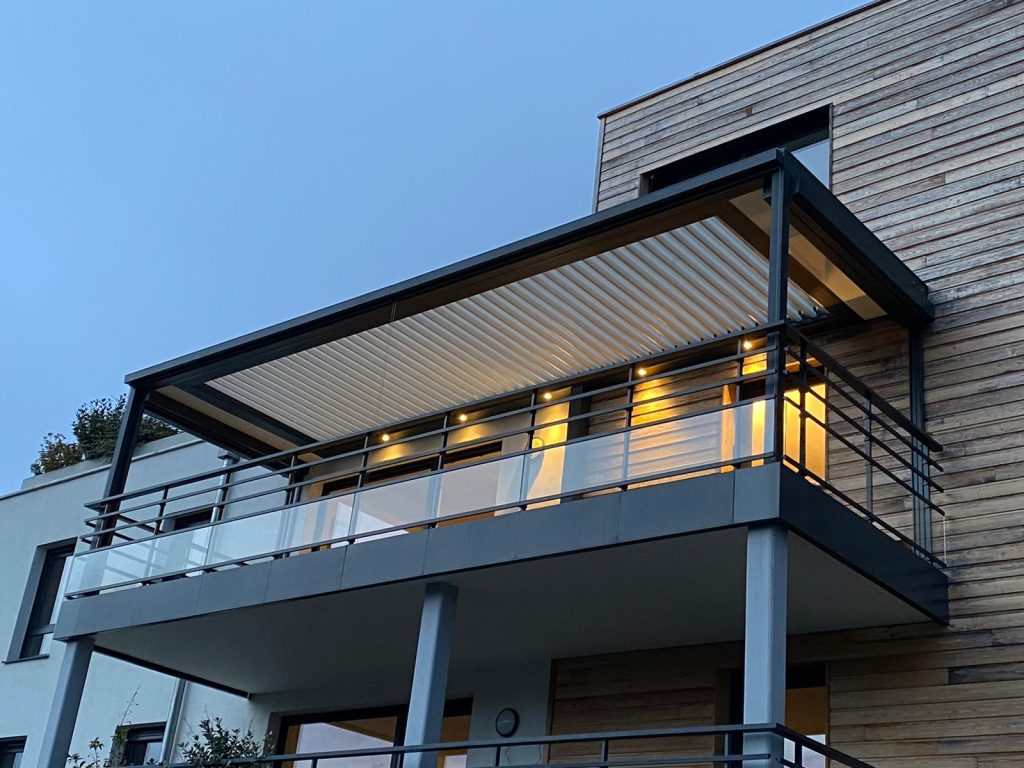 Un balcon à vivre, même le soir Pergola bioclimatique anthracite installée sur un balcon de maison à façade en bois, éclairée le soir