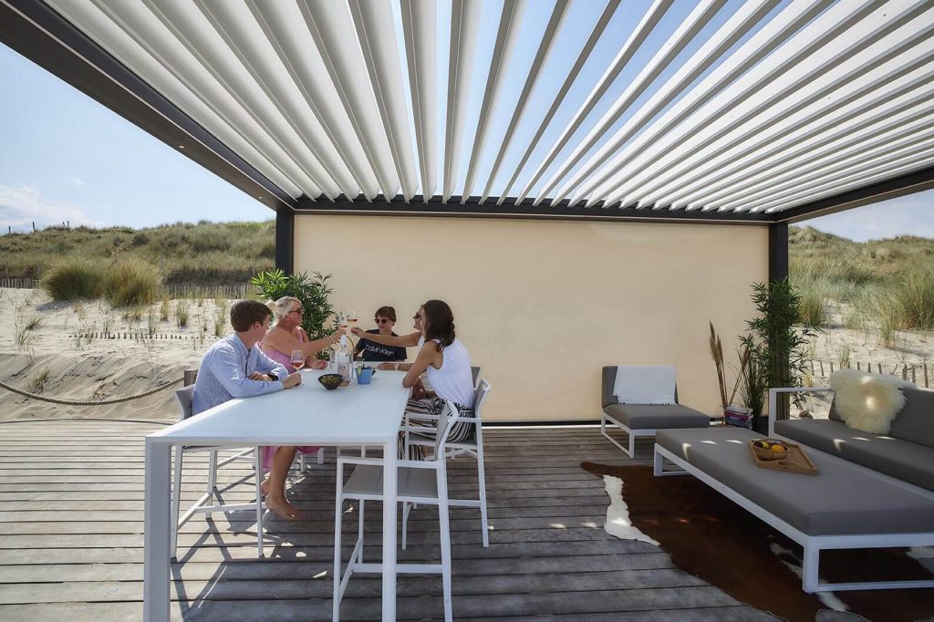 Une terrasse de plage pour se retrouver Pergola bioclimatique blanche abritant une grande table et un salon bas sur une terrasse en bois, face aux dunes