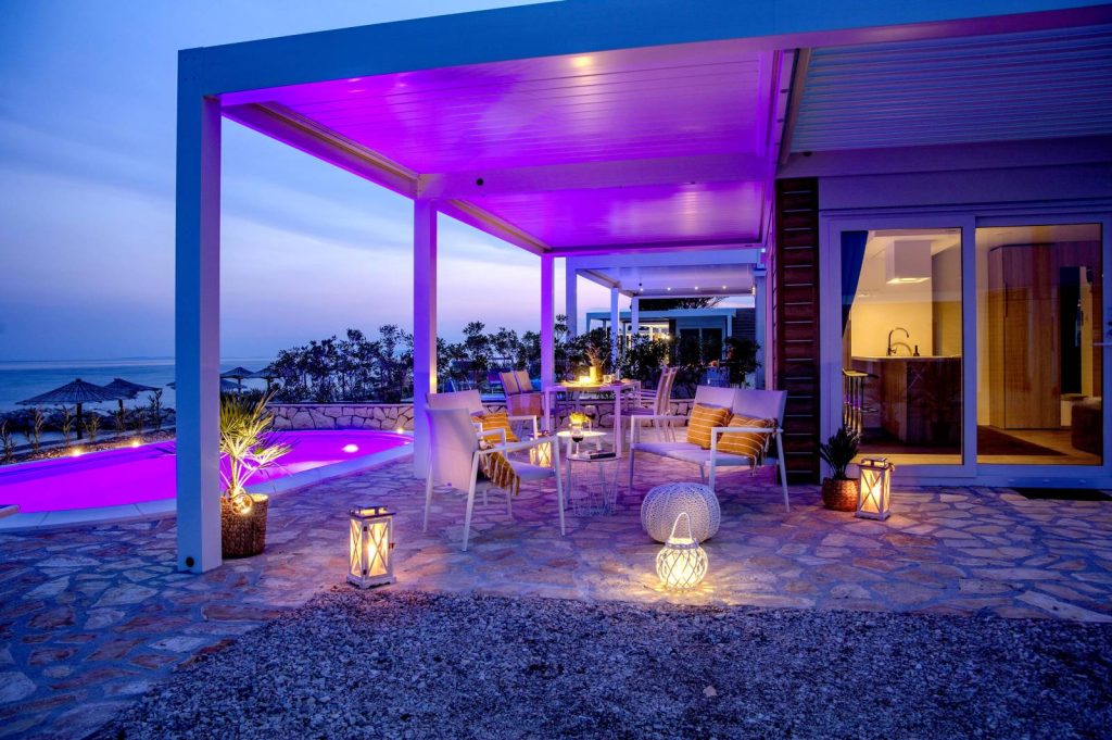 Des soirées au bord de la piscine Pergola blanche éclairée par des LED colorées au crépuscule, à côté d'une piscine