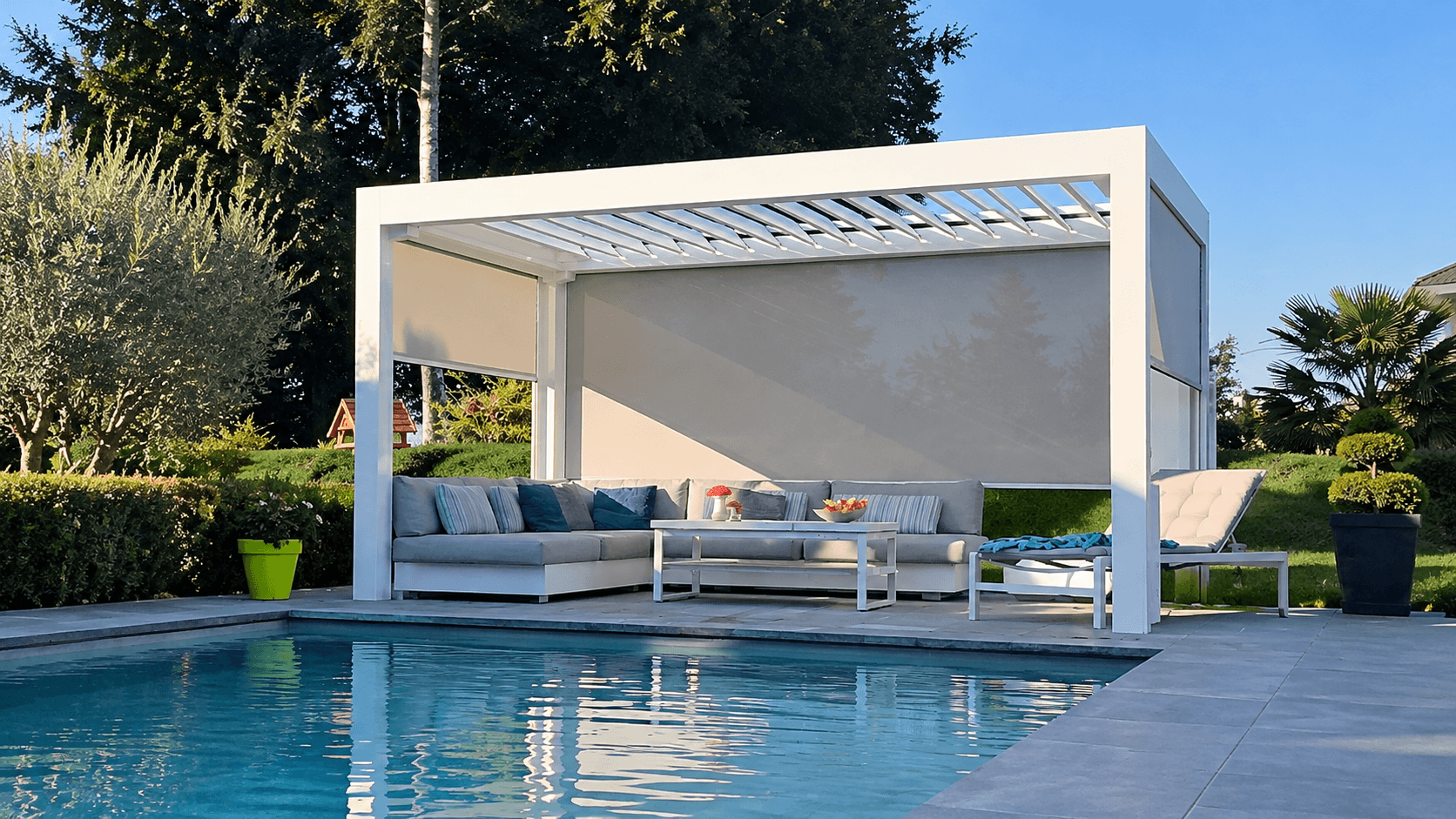 Une pergola blanche avec stores au bord de la piscine Pergola bioclimatique blanche au bord d'une piscine, stores Screen ZIP baissés à l'arrière, au-dessus d'un salon de jardin