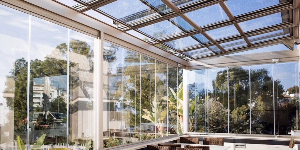 Une pergola entièrement vitrée Intérieur d'une pergola vitrée à toit en verre coulissant et parois en verre, ouverte sur des arbres