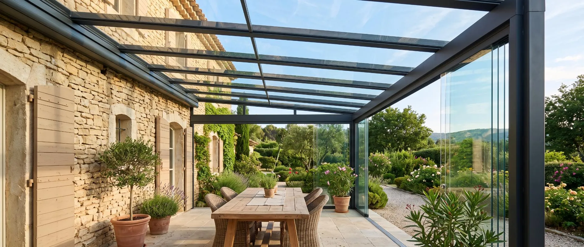 Pergola vitrée en aluminium anthracite adossée à une maison en pierre, toit en verre et rideaux de verre, table de jardin
