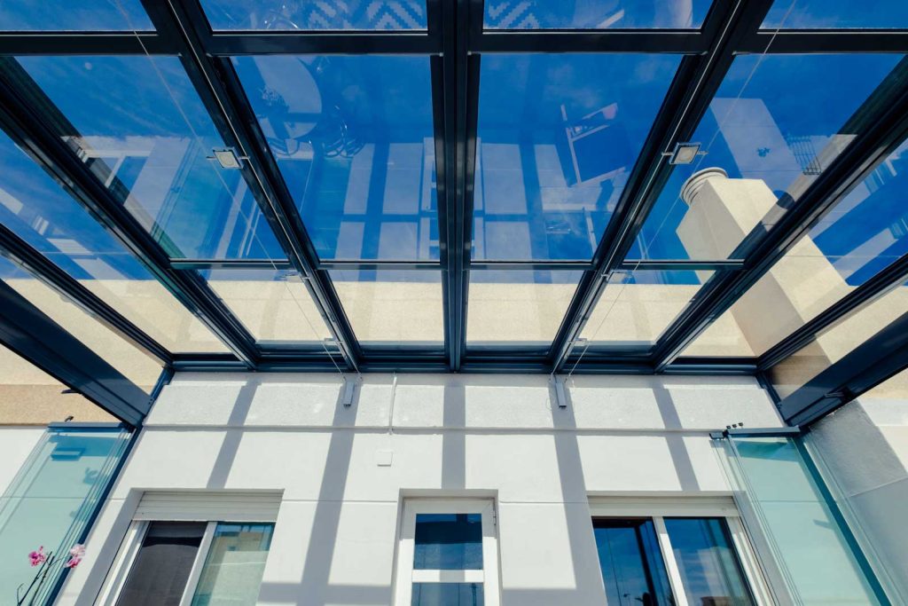 Une toiture vitrée baignée de lumière Toiture vitrée à structure aluminium gris anthracite vue de dessous, devant une façade blanche, sous un ciel bleu