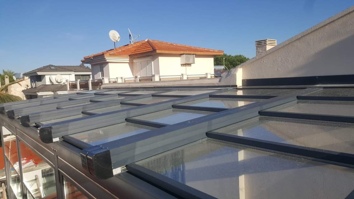 Panneaux d'une toiture vitrée rétractable anthracite installée sur un toit-terrasse, avec des maisons en arrière-plan
