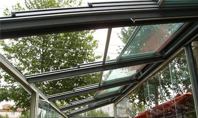 Un toit vitré qui s'ouvre sur le ciel Vue de dessous d'une toiture vitrée rétractable à profilés anthracite, avec des arbres en arrière-plan