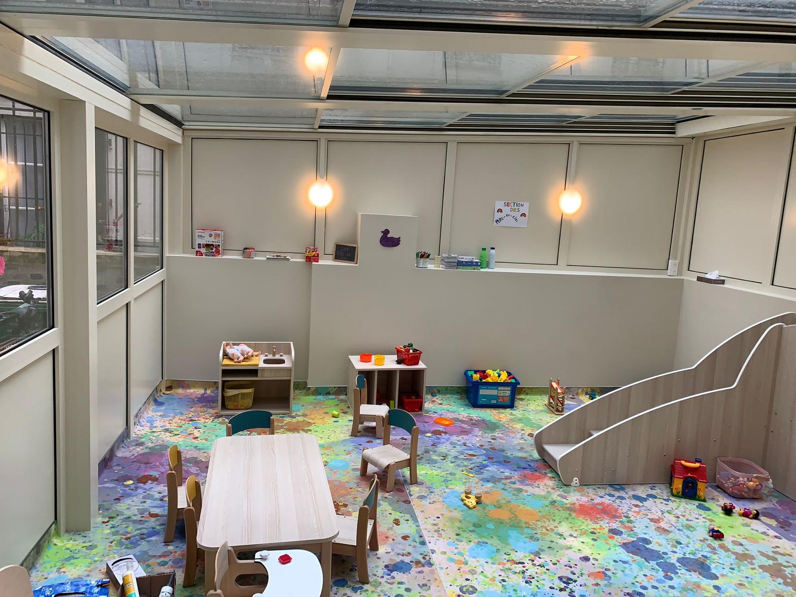 Pergola à toit vitré aménagée en espace de jeux pour enfants, avec petites tables, jouets et toboggan