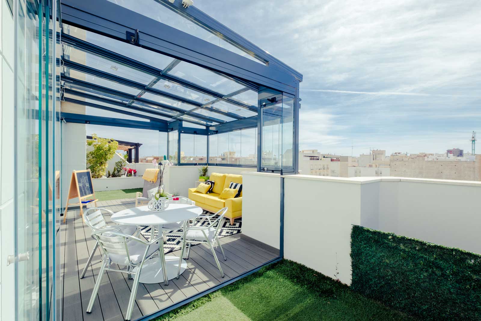 Pergola vitrée à profilés bleu foncé ouverte sur un toit-terrasse, vue de l'extérieur avec table et fauteuils